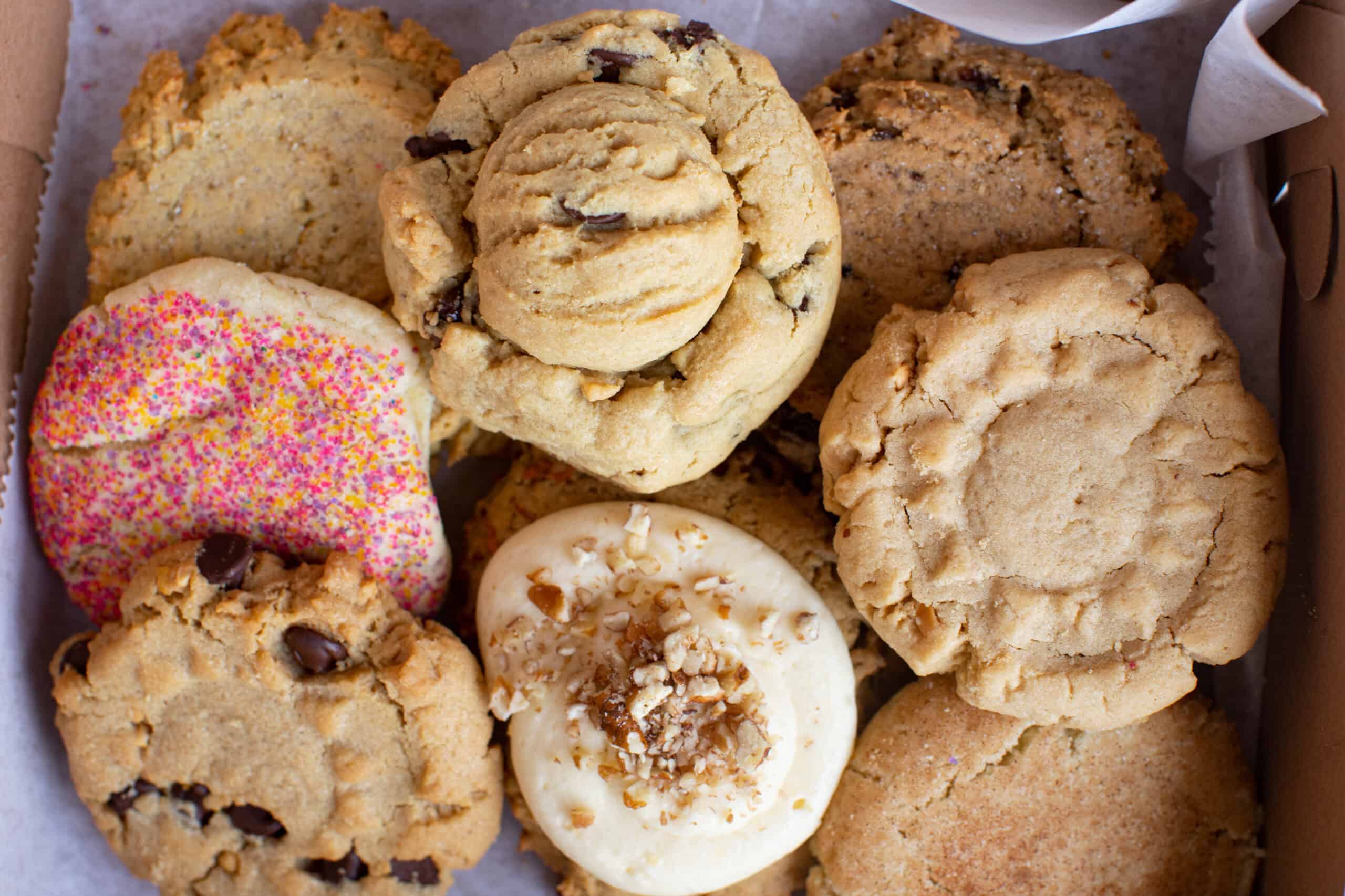 Assorted fresh-baked cookies in a box featuring colorful sprinkle sugar cookies, chocolate chip cookies, and a specialty frosted cookie. Each shows Grandma's signature hand-scooped texture.