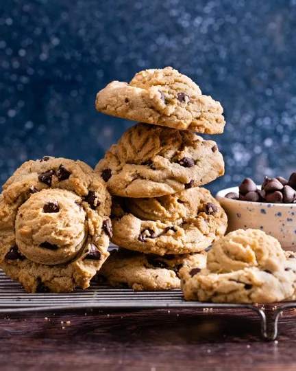 Stack of golden-brown chocolate chip cookies on dark wood, showing signature crispy edges and soft centers. Fresh-baked cookies arranged with chocolate chips scattered nearby.