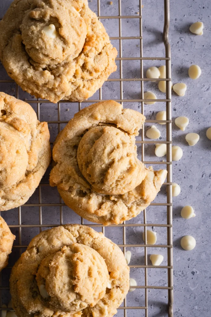 White chocolate chip cookies cooling on wire rack, showing distinctive hand-scooped texture. Close-up highlights golden-brown color and scattered white chocolate chips.