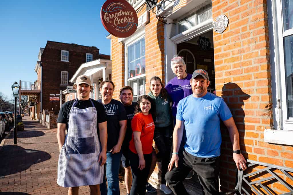 The friendly team of Grandma’s Cookies, a family-owned bakery since 1976, standing proudly in front of their Main Street shop in St. Charles, Missouri. The staff, wearing branded aprons, embodies the welcoming atmosphere and rich tradition of this beloved local establishment.